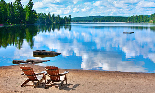 Two Muskoka chairs on a beach in front of a beautiful blue lake